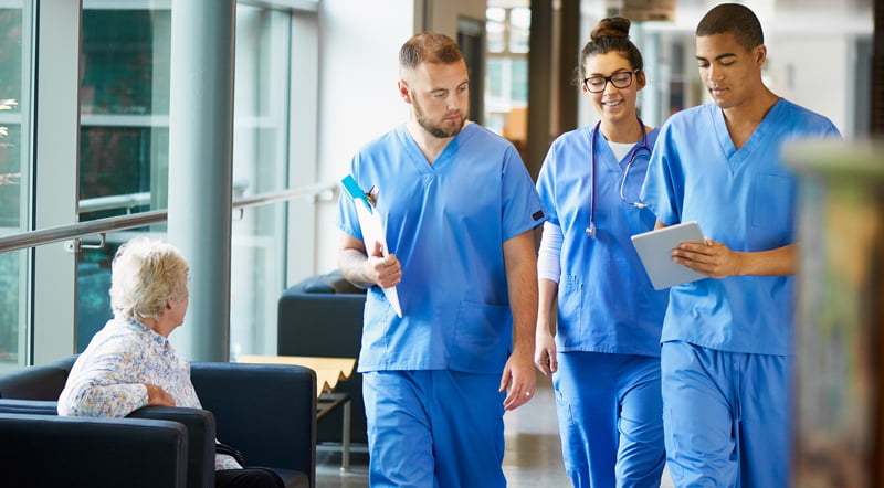 Three junior doctors wearing blue scrubs walk down a hospital corridor discussing a patient's medical case.