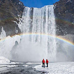 View of a couple in red anoraks dwarfed by a large waterfall with a rainbow shimmering through it, in Skogafoss, Iceland.